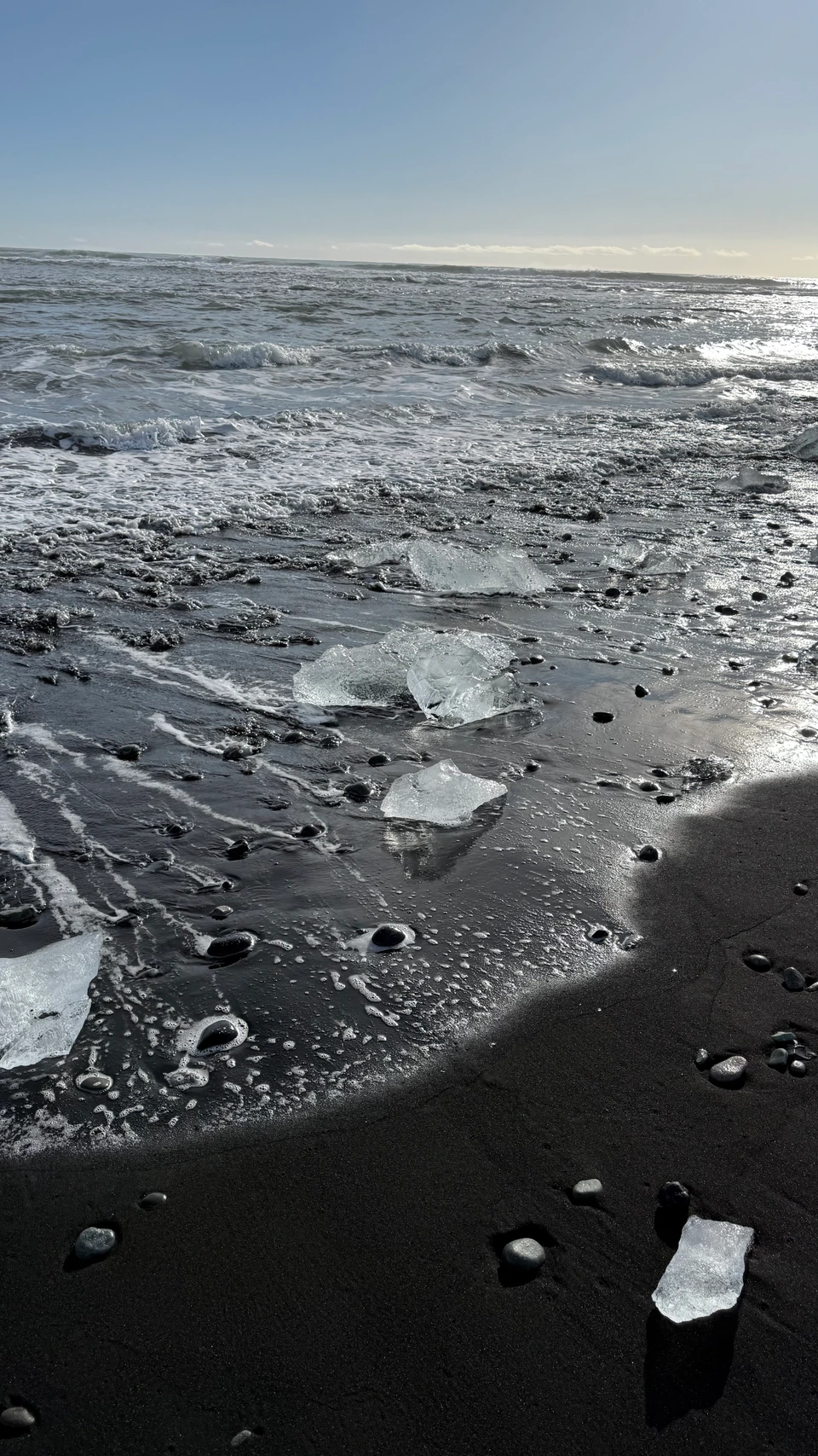 Clear ice scattered across the black sand at Diamond Beach, with waves and sunlight beyond.